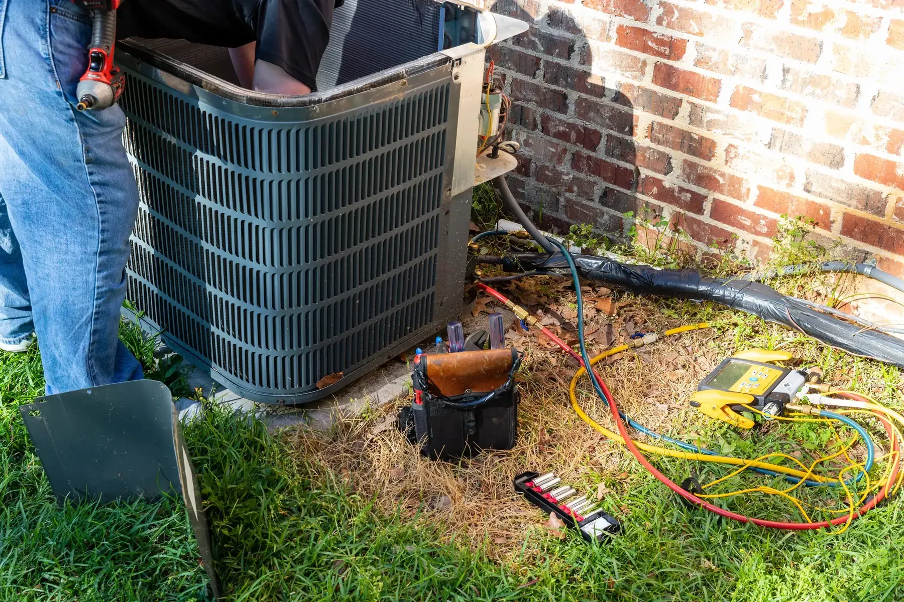 HVAC technician with the access panel removed from an outdoor condenser, inspecting the fan and wiring with tools laid out