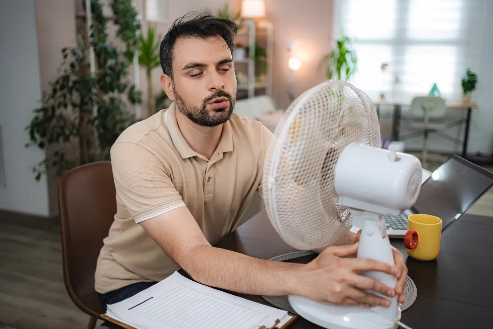 Person wiping sweat from their forehead in a hot room next to a wall thermostat during a summer heat wave